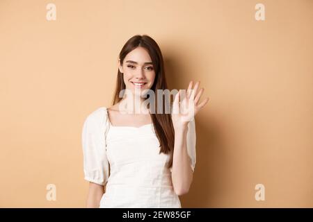 Sympathique femme souriante dire bonjour, sans avoir la main pour vous accueillir, debout gai sur fond beige Banque D'Images