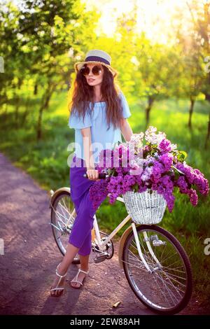 Portrait d'une jeune fille heureuse avec vélo vintage et fleurs sur fond de ville en plein soleil. Vélo avec panier rempli de fleurs. Concept de loisirs actifs. Banque D'Images