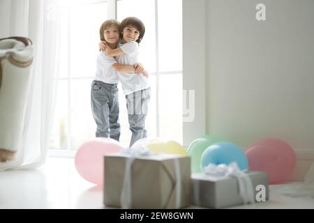 Deux adorables garçons latins jumeaux, petits enfants dans une tenue décontractée se tenant les uns les autres, regardant heureux, debout dans la chambre avec des ballons colorés et des boîtes-cadeaux. Vacances, cadeaux, concept d'enfance Banque D'Images