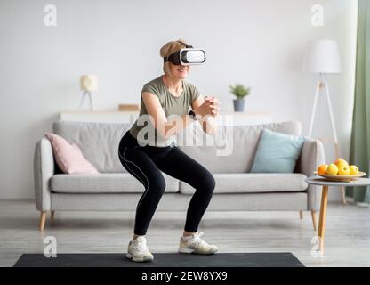 S'exercer avec le concept de réalité virtuelle. Femme athlétique mature dans un casque VR faisant des squats lors de son entraînement à la maison Banque D'Images
