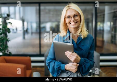 Joyeuse blonde-cheveux d'affaires femme dans les lunettes regarde la caméra, tenant l'ordinateur portable dans les bras et sourire. Belle femme d'âge moyen dans des supports d'usure élégants dans un bureau moderne Banque D'Images