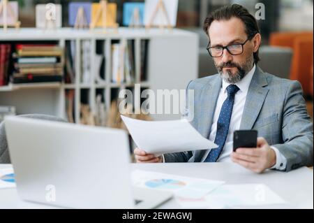 Un homme d'affaires barbu senior concentré dans des lunettes utilise un smartphone, discutant avec des collègues. Homme d'affaires sérieux assis à la réception, travaillant sur le projet importnat Banque D'Images