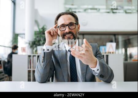 Joyeux Manager masculin aux cheveux gris sur la vidéo conférence avec des collègues, discutant du projet d'entreprise. Homme d'affaires d'âge moyen réussi souriant, regarde directement l'appareil photo, gestimant avec la main Banque D'Images