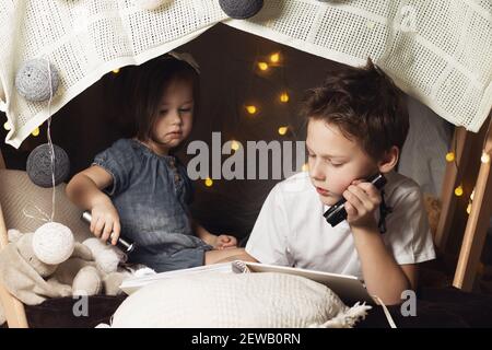 Les frères et sœurs sont dans une cabane de chaises et de couvertures. Frère et sœur, livre de lecture avec une lampe de poche à la maison Banque D'Images