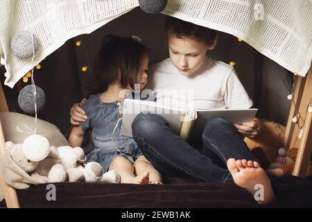 Les frères et sœurs s'assoient dans une hutte de chaises et de couvertures. Frère et sœur, livre de lecture avec lampe de poche à la maison Banque D'Images