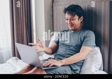 Un homme asiatique heureux travaille avec son ordinateur portable sur son lit. Concept de freelance réussi style de vie. Banque D'Images