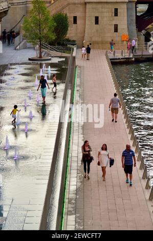 Les gens se déplacent tranquillement le long de la Chicago Riverwalk tandis que les enfants et les parents se précipitent dans la fontaine active le long de la passerelle. Banque D'Images