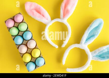 Concept de chasse aux œufs. Pâques appartement de vacances. Œufs de Pâques colorés dans un carton d'œufs et des oreilles de lapin sur fond jaune. Banque D'Images