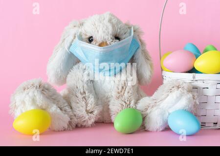 Lapin en peluche de Pâques dans un masque facial protecteur et œufs de couleur sur fond rose. Vacances d'ester pendant le concept de pandémie de coronavirus. Banque D'Images