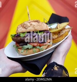 Crop anonyme personne montrant la plaque avec le hamburger acceptable avec assortiment tranches de légumes frais entre pains croquants sur fond coloré Banque D'Images