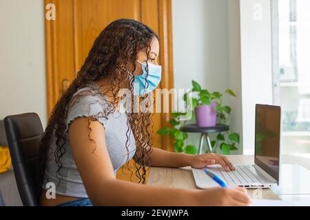 fille avec un masque sur son visage en utilisant l'ordinateur et en prenant des notes tout en télétravailleur de la maison. Banque D'Images