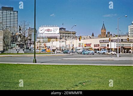Centre-ville de Denver, Colorado, États-Unis c. 1960. Cette vue de Civic Central Park est l'intersection de West Colfax Avenue et North Broadway. À gauche se trouve la fontaine des pionniers (ou monument des pionniers), une fontaine et une sculpture de Frederick William MacMonnies. La statue est de Kit Carson. Les voitures passent devant les petites gratte-ciel et les commerces de cette intersection. Un panneau géant Chevrolet s'élève au-dessus d'eux. Il indique qu'il est juste après 15h00. Le bâtiment du YMCA central est visible en arrière-plan. Aujourd'hui, le YMCA est masqué par le One Civic Center Plaza, un bâtiment de 22 étages construit là où se trouvaient les boutiques. Banque D'Images
