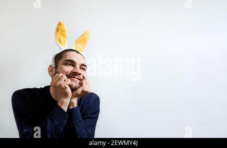 Beau caucasien avec barbe et oreilles de lièvre regarde loin et sourit avec son visage soutenu dans les mains. Créateur Pâques blanc arrière-plan homme avec yel Banque D'Images