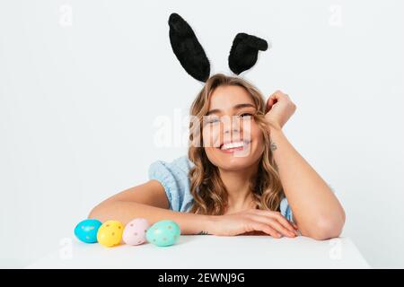 Bonne jeune femme portant des oreilles de lapin de Pâques assis à la table à côté des œufs de couleur sur fond blanc. Banque D'Images