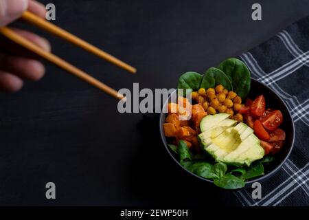 Manger avec des baguettes avocat vegan buddha bol avec tomates cerises et épinards.vue en gros plan de la nourriture maison saine sur fond sombre. Banque D'Images