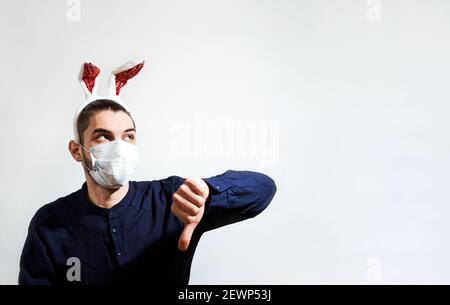 L'homme caucasien avec des oreilles de lièvre rouge porte un masque de protection médical et met sur dislike avec une main. Fêtez Pâques en vous-même. Homme avec des oreilles de Banque D'Images