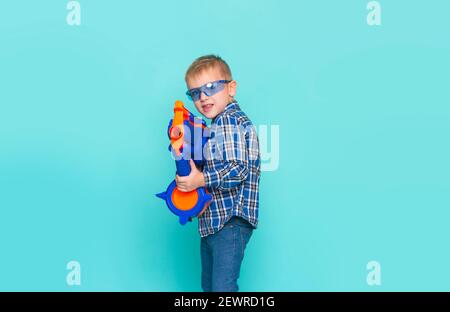 Portrait d'un enfant jouant avec un pistolet en plastique, isolé sur fond bleu. Magnifique adolescent de race blanche s'amuser avec un jouet pour enfants Banque D'Images