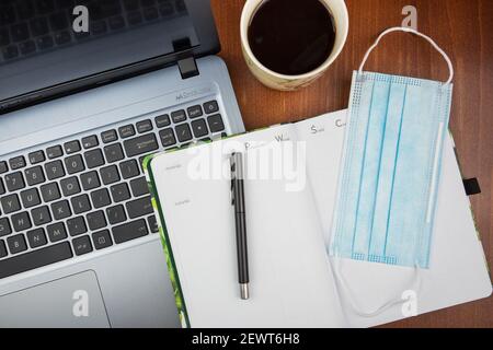 KATOWICE, POLOGNE - 12 février 2021 : vue de dessus d'un bureau avec ordinateur portable. Un stylo noir et un masque chirurgical allongé sur un planificateur quotidien. Une tasse de café à côté. Banque D'Images