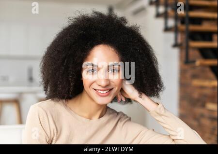 Portrait en tête de jeune femme indépendante afro-américaine avec une coiffure frino-curly et un beau sourire, placé la main sur la joue, regardant l'appareil photo tout en étant debout dans la salle de séjour Banque D'Images