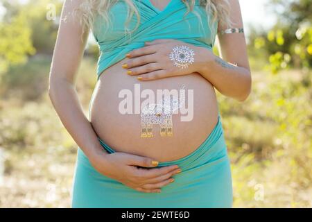 Ventre de fille blonde enceinte avec tatouage flash d'argent temporaire, symbole éléphant, en attente de bébé garçon, en tenue bleue, à l'arrière-plan de la nature Banque D'Images