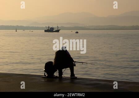 Un homme assis pêche avec de petits bateaux et un chalutier retournant au port après une nuit de pêche silhoueté Santander Bay Cantabria Espagne hiver Banque D'Images