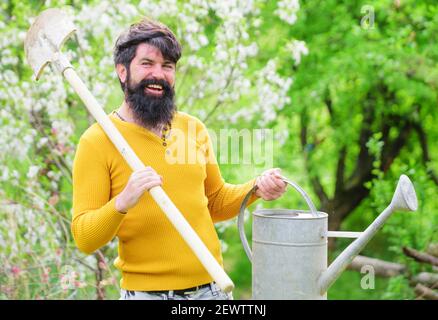 Ressort. Jardinier barbu avec arrosoir et bêche. Homme souriant se préparant à planter. Arrosage. Agriculteur travaillant dans le jardin. Banque D'Images