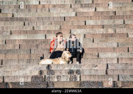 Un jeune couple avec un chien assis sur les marches de la cathédrale d'Helsinki lors d'une chaude et ensoleillée journée d'hiver à Helsinki, en Finlande Banque D'Images