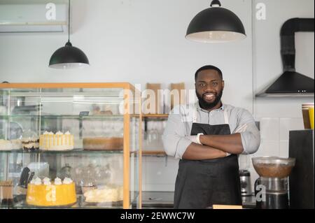 Souriant et sympathique propriétaire de petite entreprise afro-américaine, gérant de café, serveur de boulangerie multiracial se tient derrière le comptoir dans une posture confiante avec les bras croisés et regarde la caméra Banque D'Images