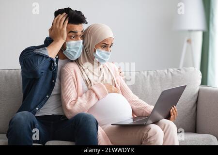 Couple musulman enceinte choqué dans des masques médicaux assis avec un ordinateur portable À la maison Banque D'Images