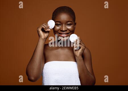 Soin de la peau. Portrait d'une femme africaine joyeuse tenant deux tampons de coton, visage nettoyant Banque D'Images