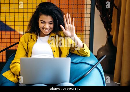 Une jeune femme afro-américaine joyeuse a des rencontres en ligne avec des collègues ou des amis. Une étudiante ou une employée heureuse utilise un ordinateur portable, est assise dans une chaise confortable, concept de communication Banque D'Images