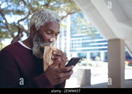 Homme afro-américain senior dans la rue en train de manger des sandwichs et en utilisant smartphone Banque D'Images