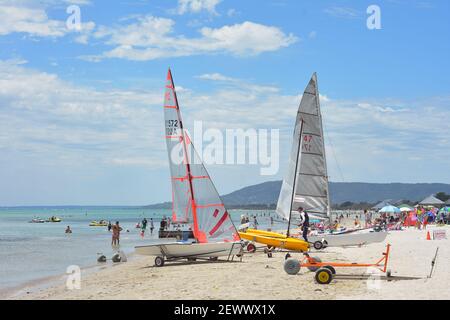 Petits bateaux à voile garés dans des remorques de plage sur la rive avec des vacanciers appréciant l'eau peu profonde ou se cachant à l'ombre des parasols en arrière-plan. Banque D'Images