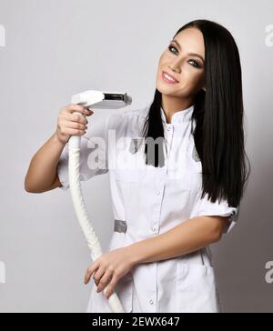 Jeune femme brune souriante médecin cosmetologue infirmière en uniforme médical montre l'enlèvement des cheveux au laser de l'équipement médical Banque D'Images