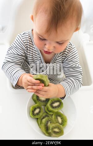 Mignonne bébé fille sont assis avec un fond blanc kiwi intérieur. Drôle d'enfant explore le fruit Banque D'Images