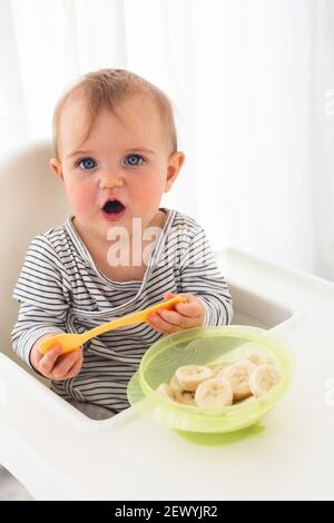 Mignonne bébé fille sont assis whith tranché banane fond blanc intérieur. Drôle d'enfant explore le fruit Banque D'Images