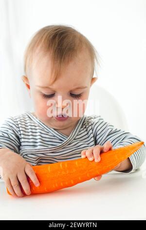 Mignonne bébé fille sont assis avec la carotte blanc fond intérieur. Drôle d'enfant explore les légumes Banque D'Images