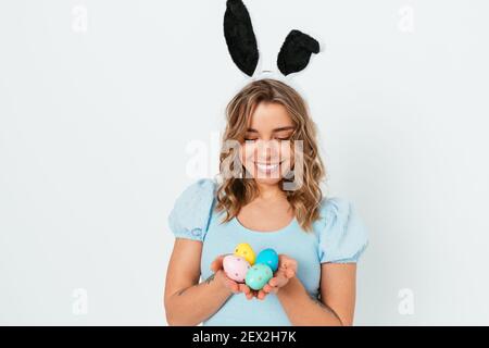 Jolie jeune femme blonde portant des oreilles de lapin de Pâques tenant des œufs de couleur dans ses mains et souriant tout en se tenant debout sur fond blanc de studio. Banque D'Images