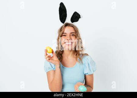 Jeune femme joyeuse portant des oreilles de lapin de Pâques tenant des œufs colorés souriant regardant l'appareil photo tout en se tenant sur fond blanc de studio. Banque D'Images