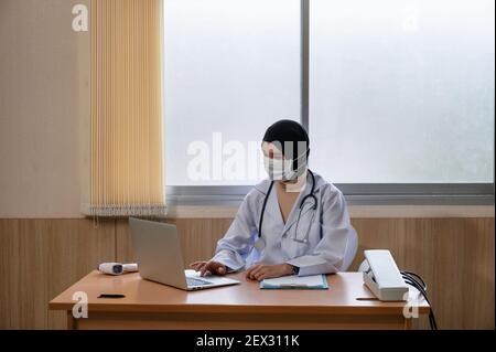 Femme musulmane médecin portant le hijab en uniforme médical à l'aide d'un ordinateur portable et d'un sphygmomanomètre, thermomètre infrarouge sur le bureau de l'hôpital Banque D'Images