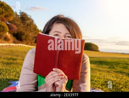 Une femme d'âge moyen se faisant un pelage sur un livre qu'elle tient, tout en étant couchée sur un peu d'herbe. Coucher de soleil crémeux en arrière-plan. Banque D'Images