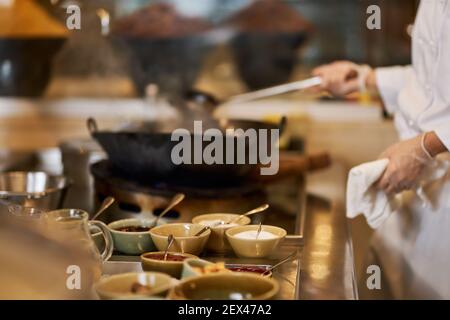 Photo rognée d'un ouvrier de cuisine debout près d'une table avec des ingrédients tout en les ajoutant dans un wok Banque D'Images