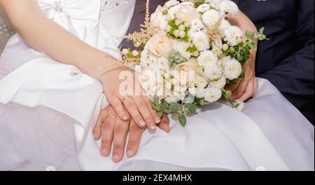 Les mains des jeunes mariés se reposent l'une sur l'autre. La paume de la mariée repose sur le dessus de la paume du marié. Il y a un bouquet de mariage à proximité. Banque D'Images