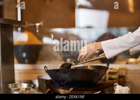 Photo rognée des mains du chef en gants ajoutant d'autres ingrédients dans un wok sur un poêle Banque D'Images