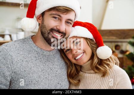 Beau couple heureux en chapeau de père noël embrassant et souriant à l'appareil photo dans la cuisine confortable Banque D'Images