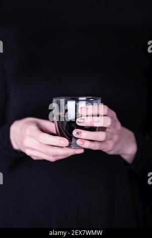 Les femmes tiennent une tasse en verre transparent de café noir. Manucure avec vernis à ongles de couleur rose, motif floral noir Banque D'Images