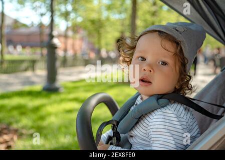 Gros plan d'un adorable tout-petit assis dans un pram et regardant l'appareil photo sur le fond du parc public vert. Banque D'Images