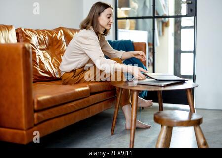 Jolie femme assise sur le canapé marron à la maison tout en prenant la tablette numérique de la table. Concept de style de vie et de travail à domicile. Banque D'Images