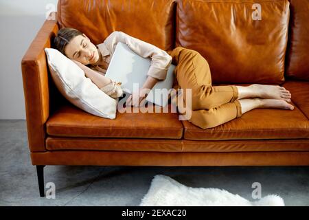 Une jolie femme s'est endormie sur le confortable canapé marron dans une étreinte avec un ordinateur portable. Concept de style de vie. Banque D'Images