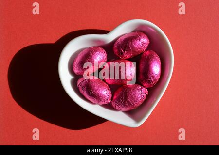 Œufs de Pâques en forme de coeur blanc, enrobés de papier d'aluminium rose plat isolé sur fond rouge Banque D'Images